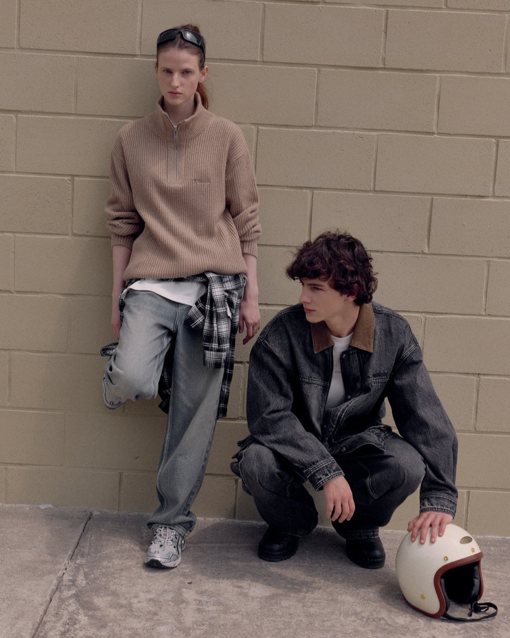 Two people standing against a beige brick wall with a helmet on the ground.