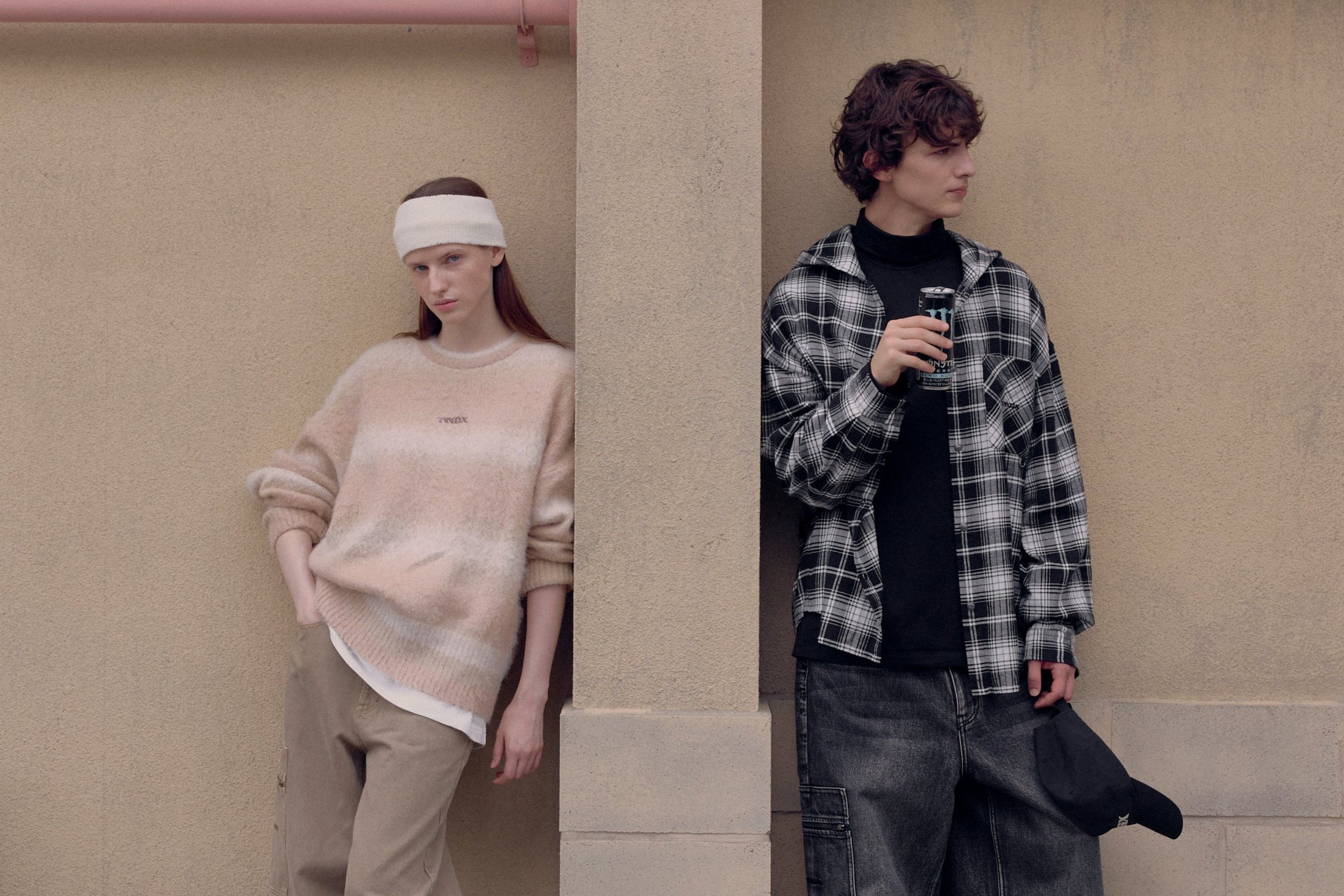 Two people standing against a beige wall, one wearing a headband and beige outfit, the other in a plaid shirt and dark pants.